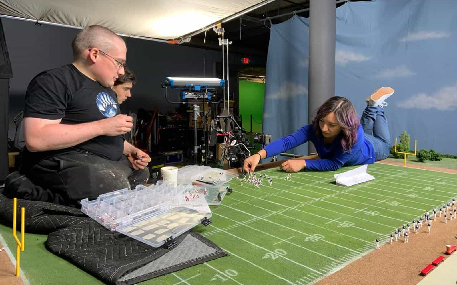 Olivia lying on her stomach adjusting diorama pieces on the "The Herricanes" set. The diorama is a miniature football field with miniature football figurines.