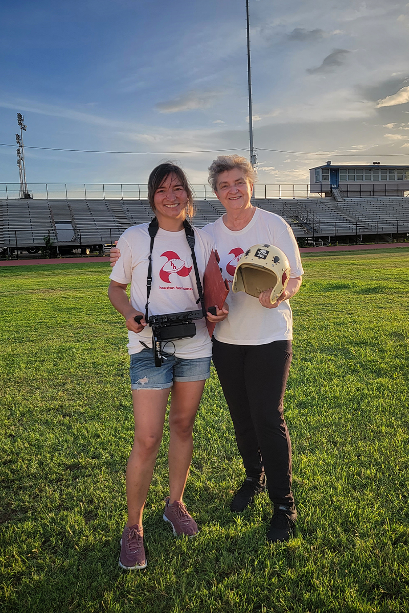 Olivia Kuan and her mother Basia Haszlakiewicz on a football field wearing matching Houston Herricanes t-shirts. Olivia has a camera around her neck and her mother is holding a football helmet.