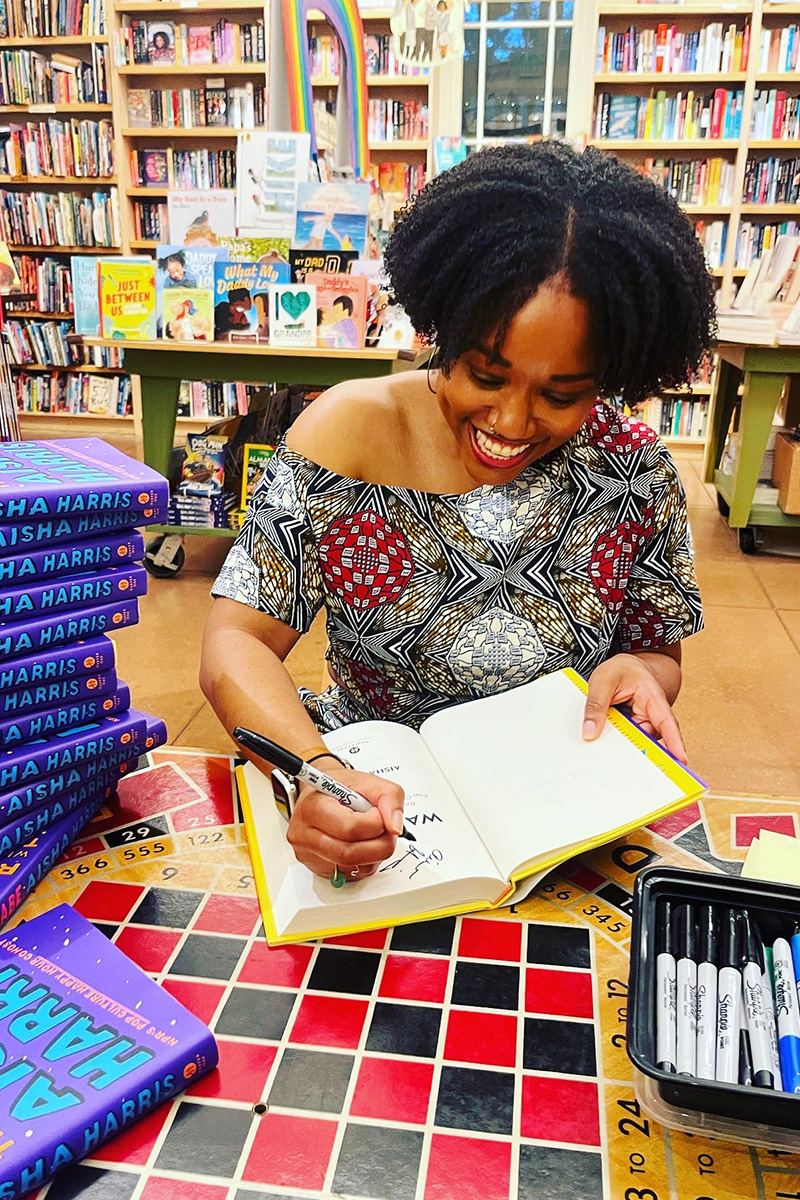 Aisha smiling and signing a book on a checkers table. There is a pile of her book, WANNABE, next to her.