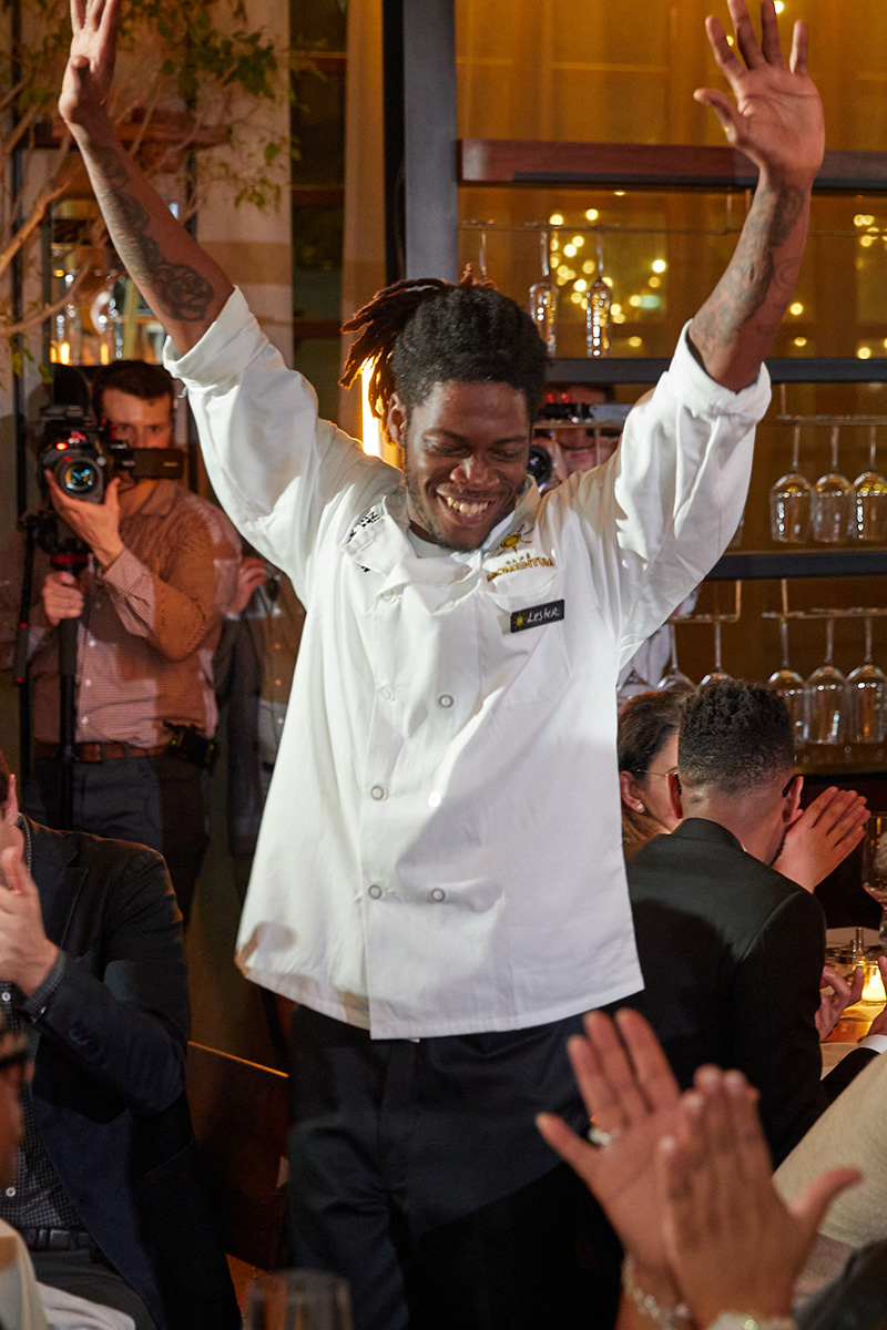 A Café Momentum intern wearing a white chef's coat, smiling, with his arms raised up in a celebratory fashion.