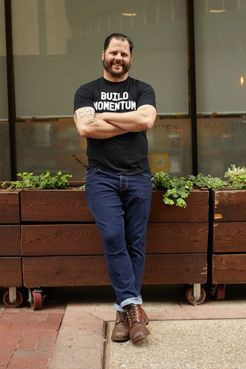 Chad House in front of Café Momentum Dallas with his arms and legs crossed, leaning up against a wooden planter. He is wearing jeans, brown shoes, and a black t-shirt that says “build momentum.”