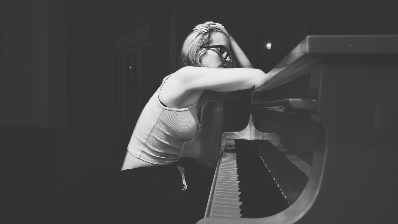 A black and white image of Ingrid sitting in front of a piano, with one arm leaning on the top of the piano and the other propping up her head.