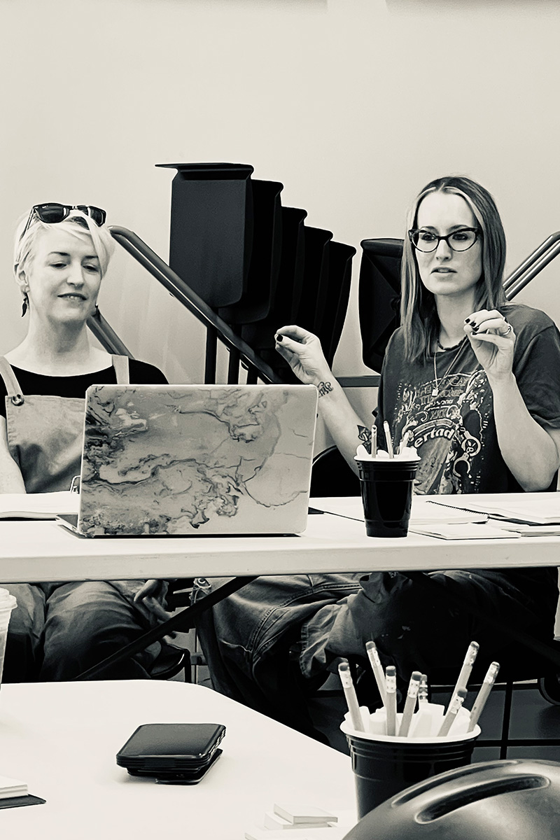 A black and white image of Ingrid Michaelson and Carmel at a table. Carmel is working on a laptop and Ingrid is gesticulating with her hands.