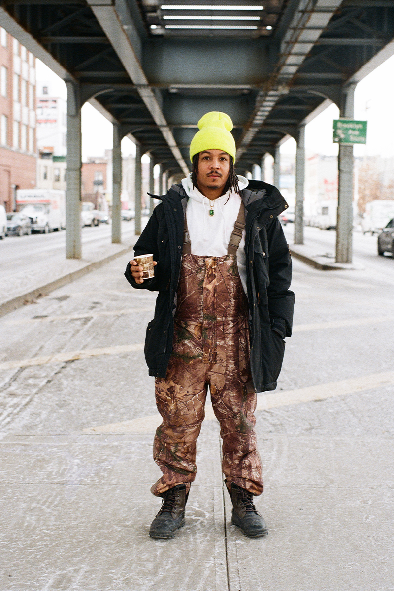 Chad Sanders under an overpass in New York. He is wearing a yellow beanie, camo overalls, a black jacket, and is holding a coffee cup.