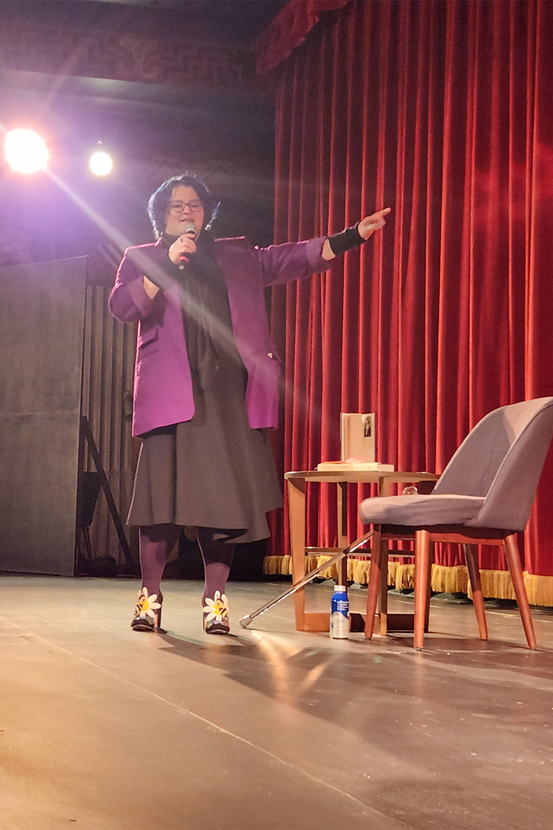 Emily on stage speaking into a mic with her left arm raised out. She is wearing a purple blazer, a gray dress, and floral heels.