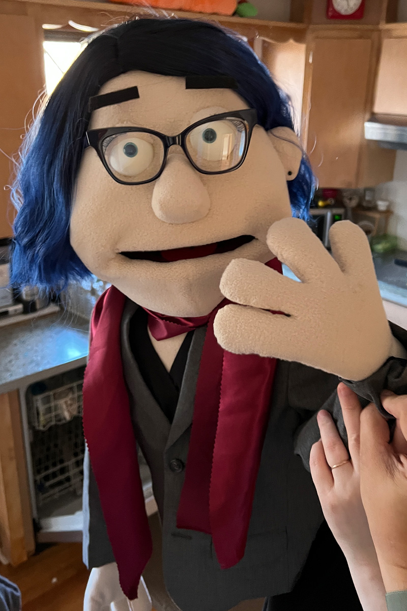 A puppet with blue hair and black frame glasses holding its hand up to its face.